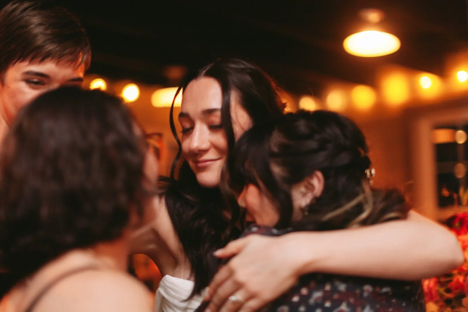 A group of friends hugging and sharing an emotional moment at a social gathering, with warm indoor lighting.