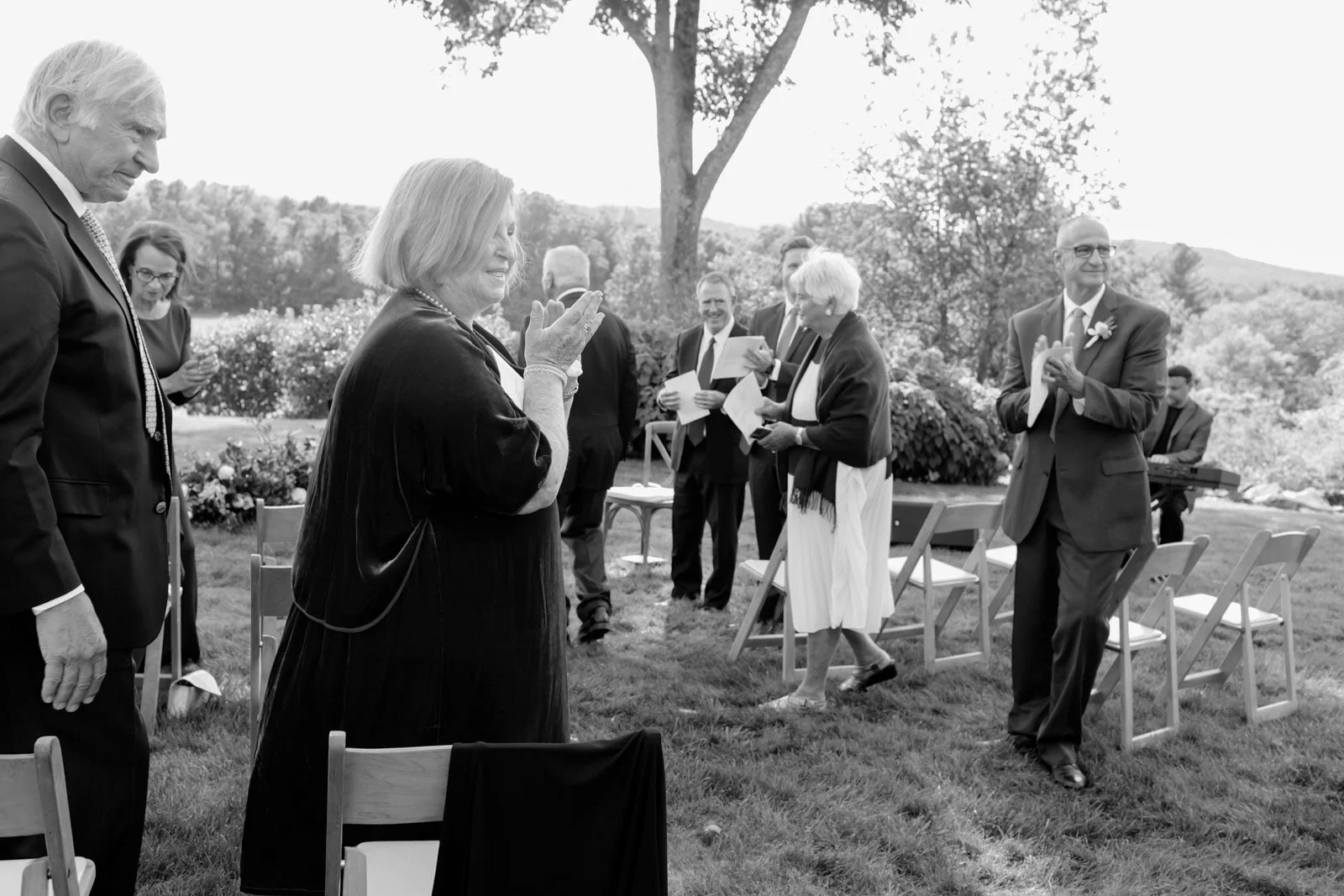 People gathered outdoors for a celebration, with some clapping and singing, and a musician playing a keyboard in the background.
