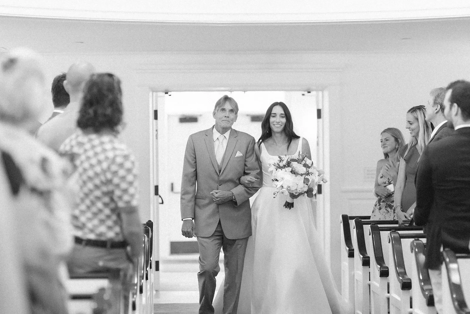 A bride walking down the aisle with her father at a wedding ceremony, holding a bouquet of flowers, with guests watching on.