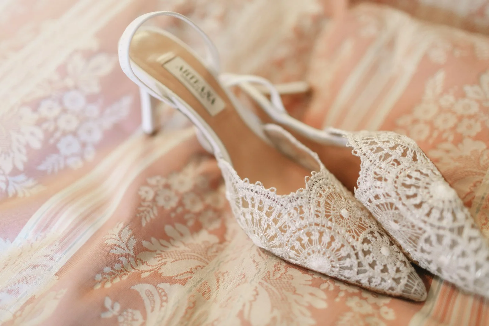 A pair of white lace wedding shoes on a peach and cream patterned fabric surface.