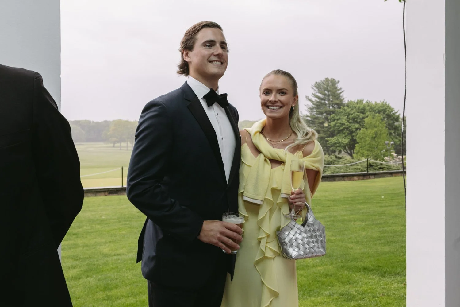 A couple dressed in formal attire, the man in a tuxedo holding a glass of beer and the woman in a yellow dress holding a glass of champagne and a silver handbag, standing outdoors at a garden or golf course.