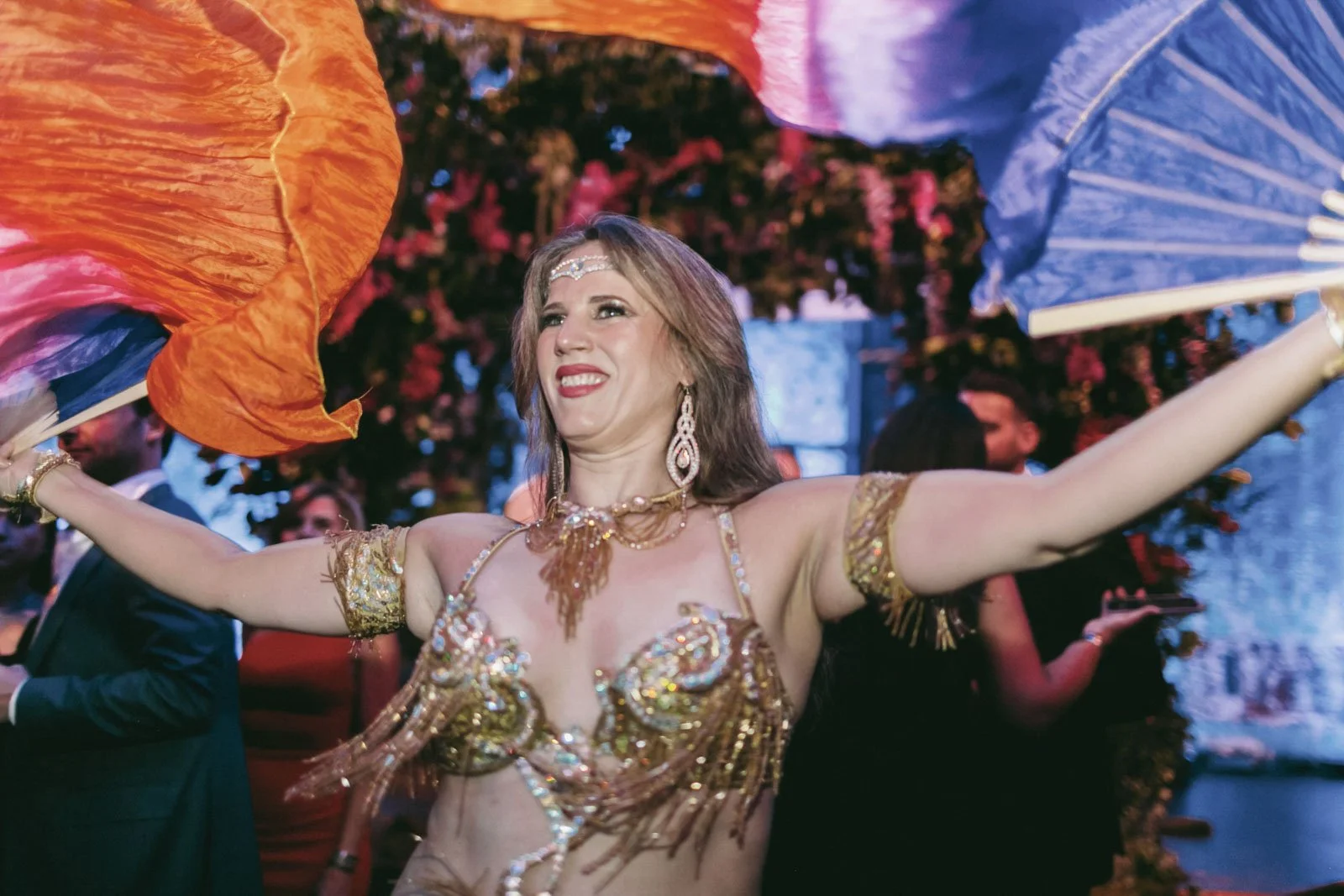 A woman dancing at a celebration, holding colorful fabric or flags with her arms outstretched, surrounded by other people, with flowers and lights in the background.