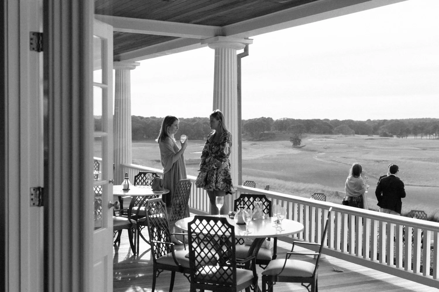People on a porch overlooking a field in the daytime, with two women standing and talking, and a couple walking away in the background.