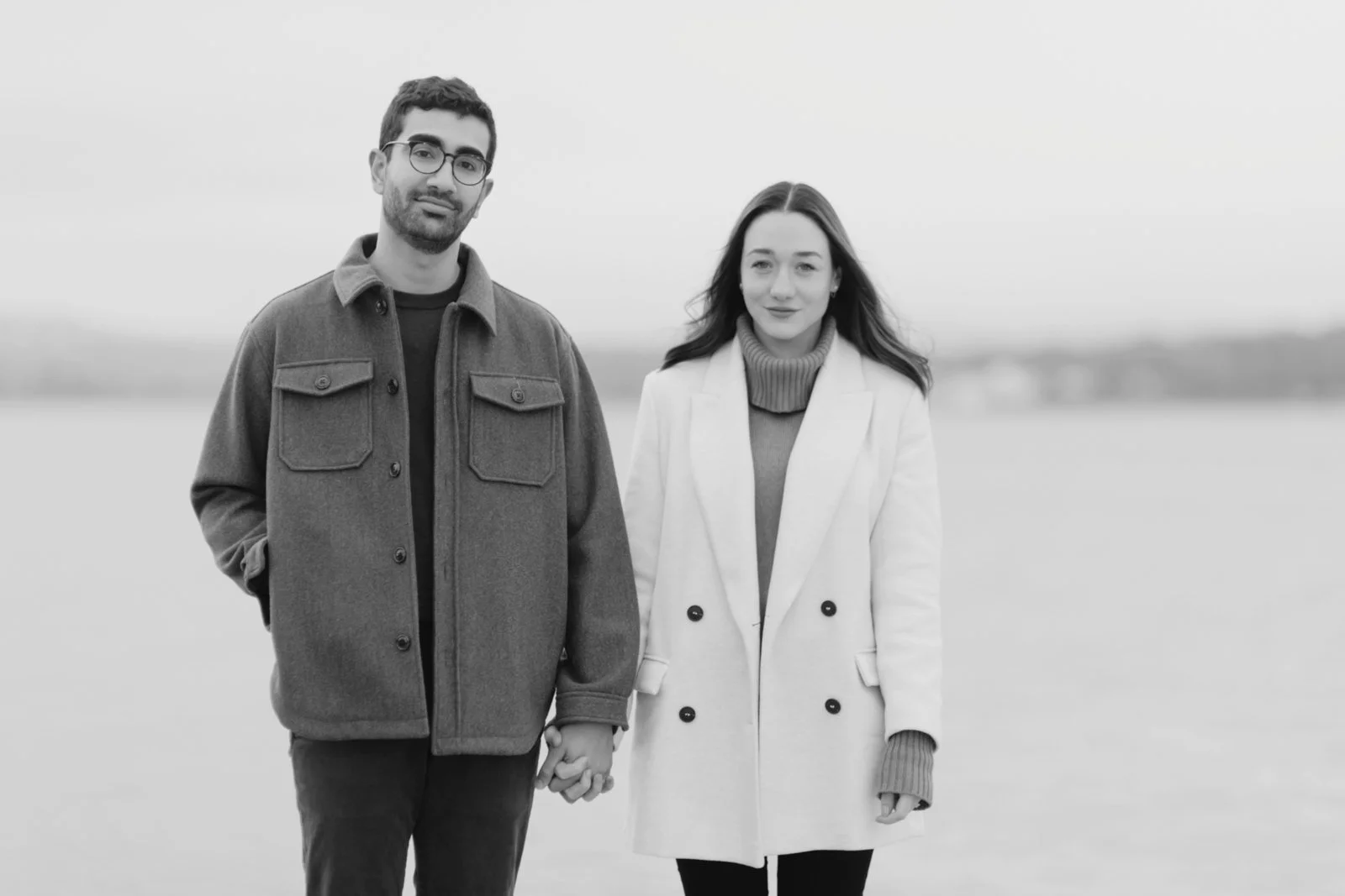 A black-and-white photo of a man and woman standing together outdoors near a body of water, holding hands, dressed in warm clothing, facing the camera.