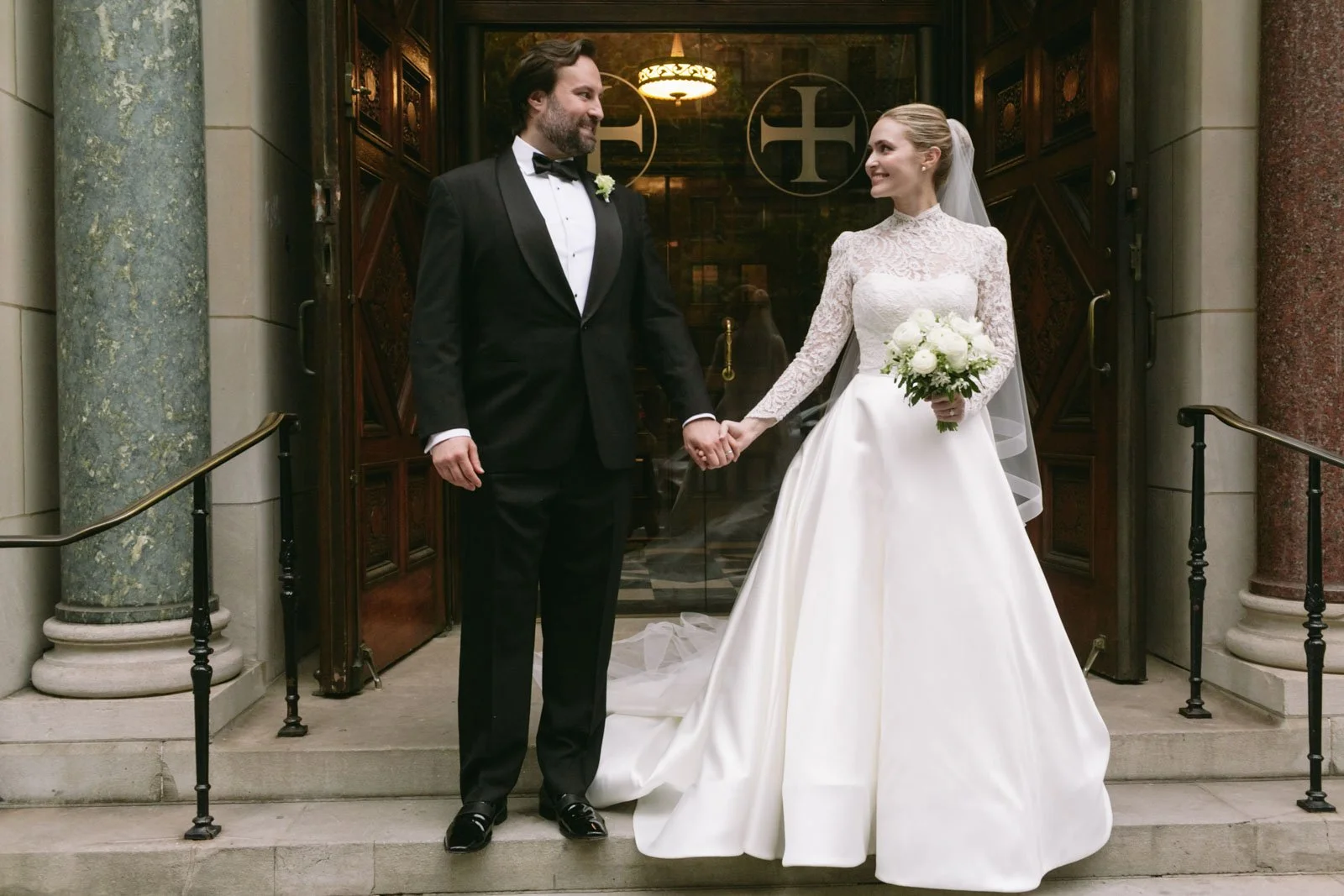 A bride and groom standing on the steps outside Christ Church NYC holding hands and smiling at each other. The bride is wearing a white lace wedding dress, holding a bouquet of white flowers, and has a veil. 