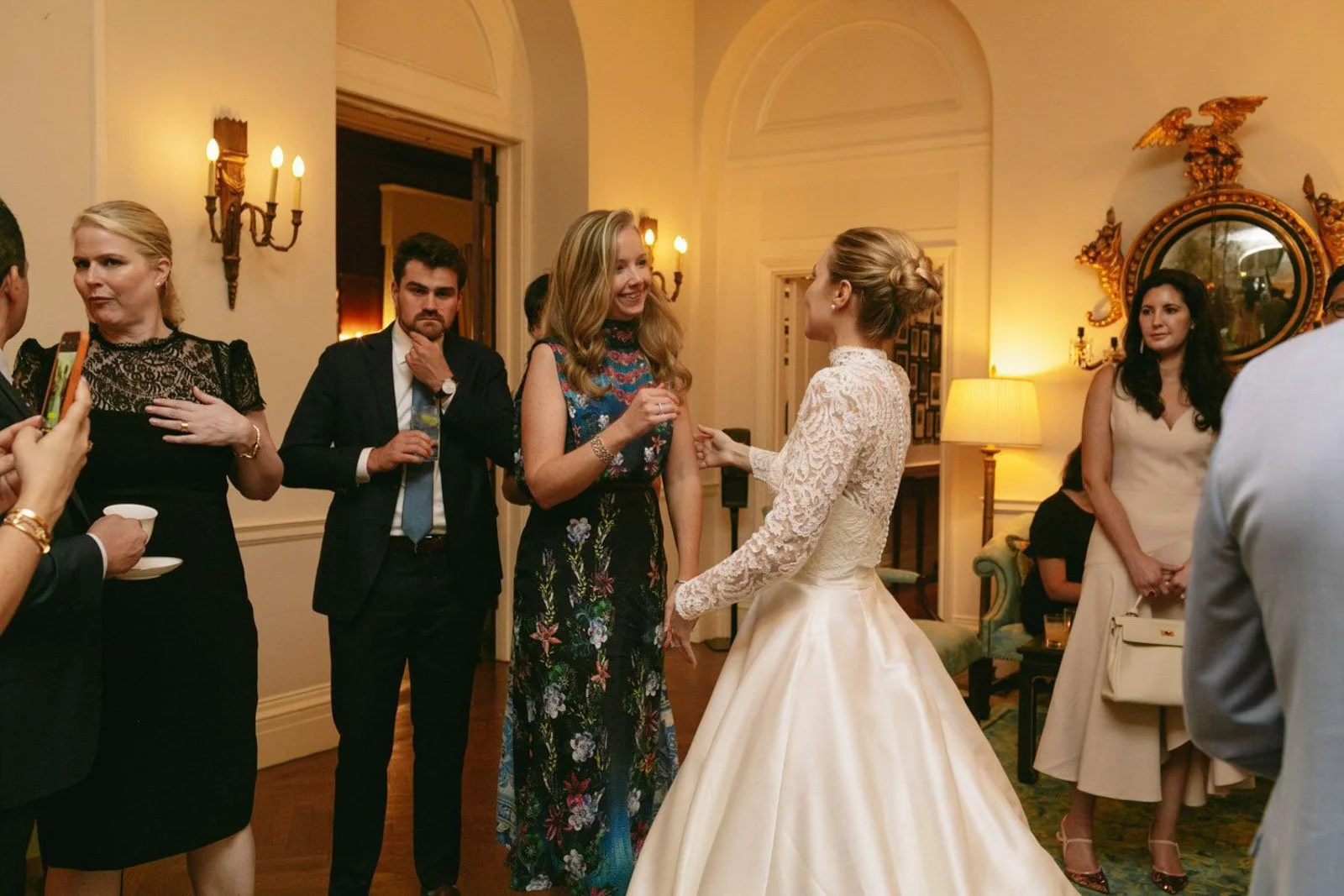 A wedding reception with women and men in formal attire, indoors with warm lighting, featuring a bride in a white wedding gown and a guest in a floral dress smiling and talking.