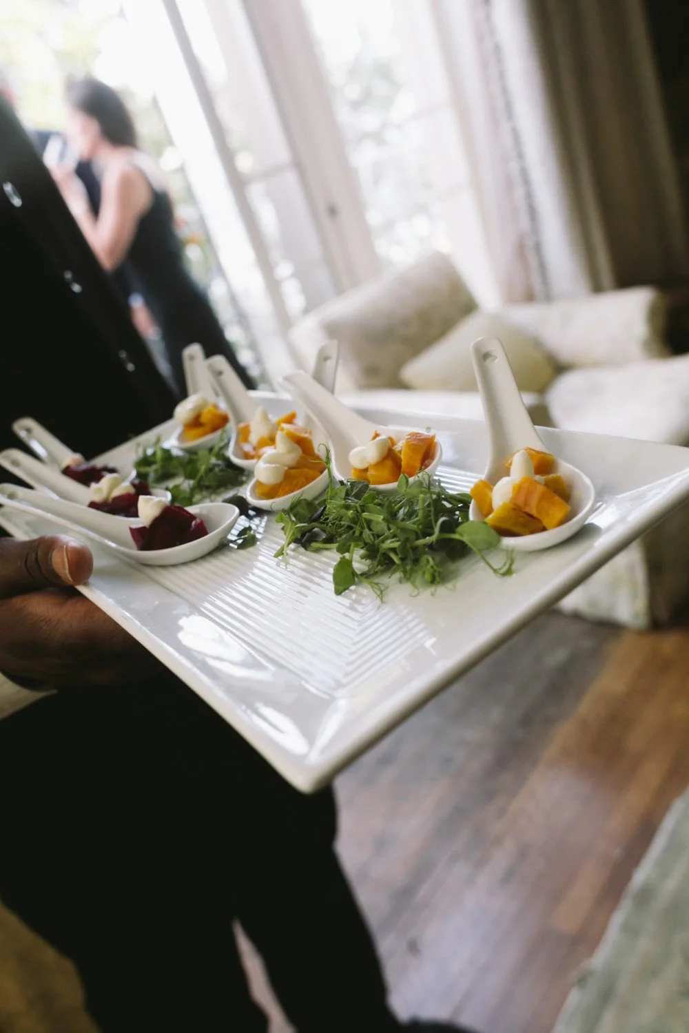 A person holding a rectangular tray with four small white porcelain spoons filled with appetizers, garnished with fresh herbs, set on a wooden table near a window with sunlight and a blurred woman in black in the background.