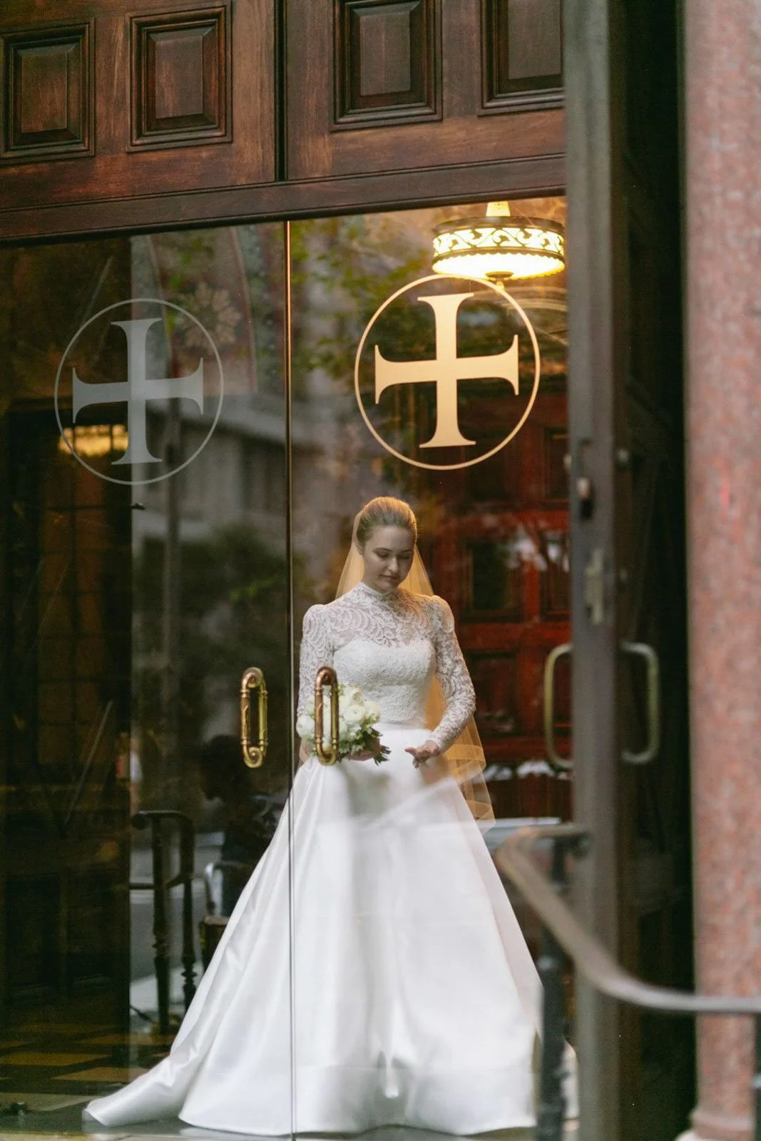 A bride in a white wedding dress with lace details, holding a bouquet, standing inside a building and looking down, as seen through glass doors with religious symbols.