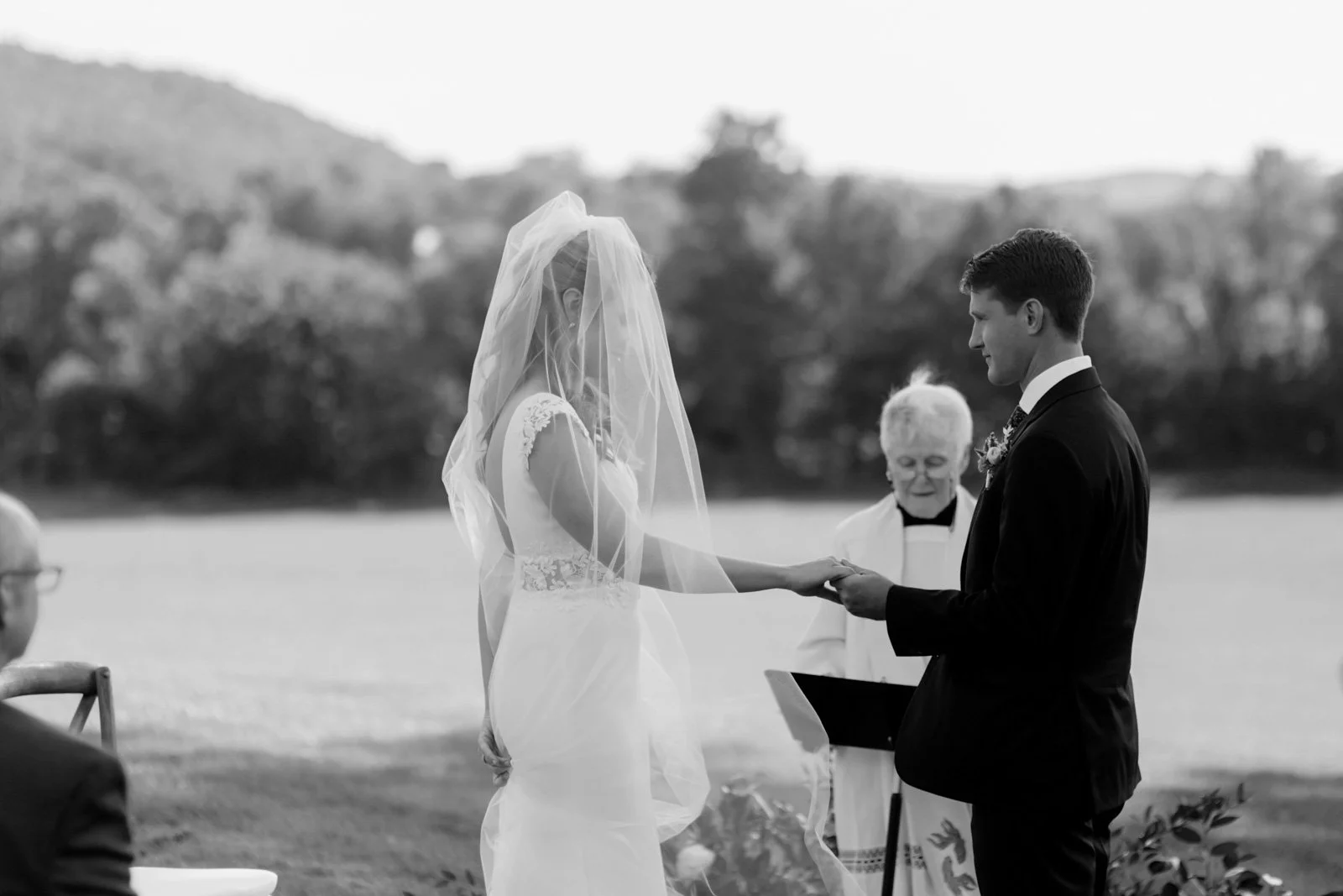 A bride and groom exchange vows outdoors, with an officiant standing behind them, by a lake and trees.