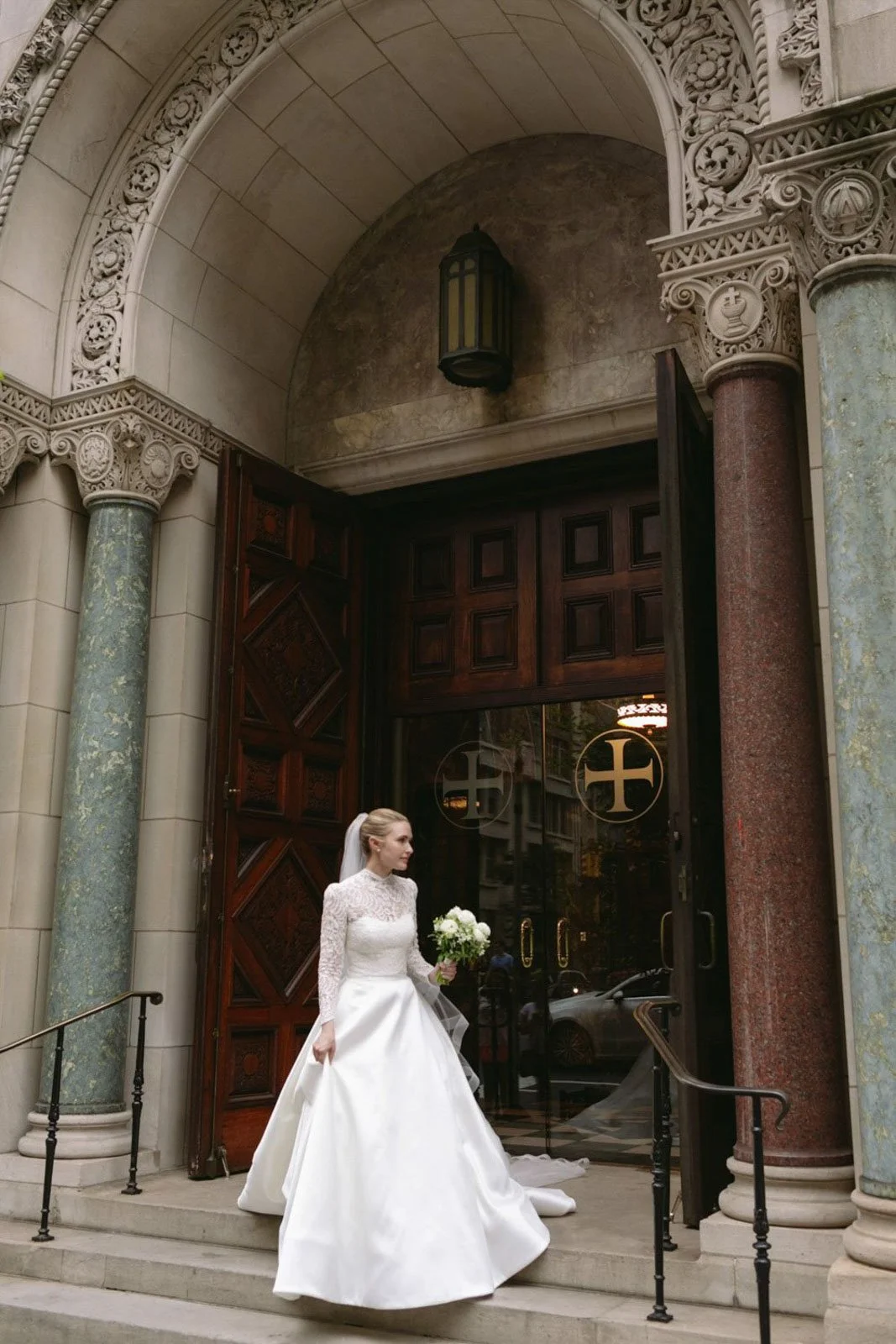 Bride in a white wedding gown holding a bouquet, standing on the steps outside Christ Church NYC with ornate columns and large wooden doors.