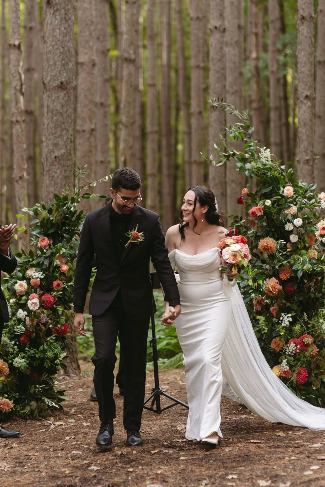 Bride and groom walking hand in hand during their wedding ceremony in a forest, surrounded by floral arrangements.