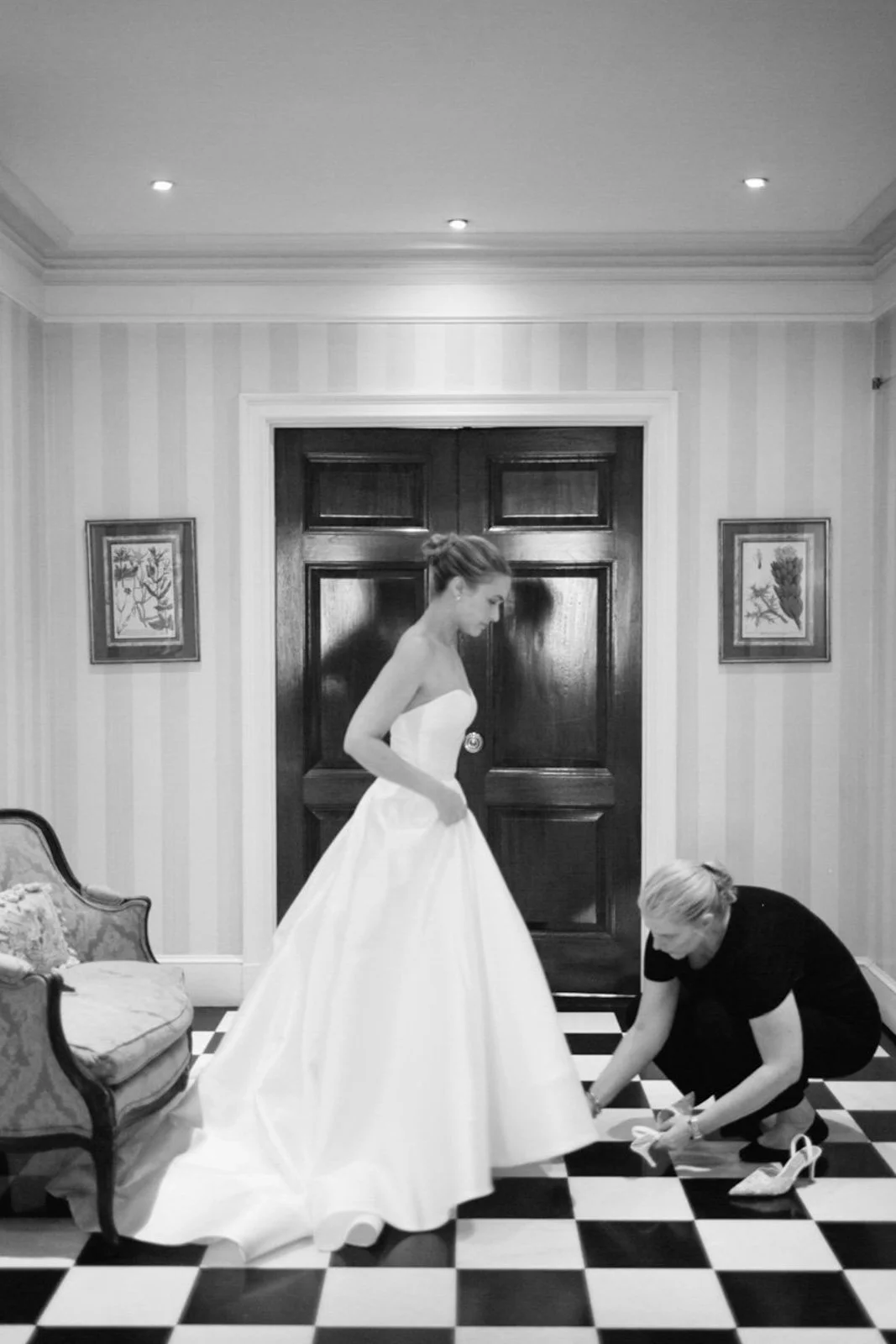 A bride in a strapless wedding gown stands as a woman helps her put on wedding shoes inside a room with striped wallpaper and framed artwork, black and white photo.
