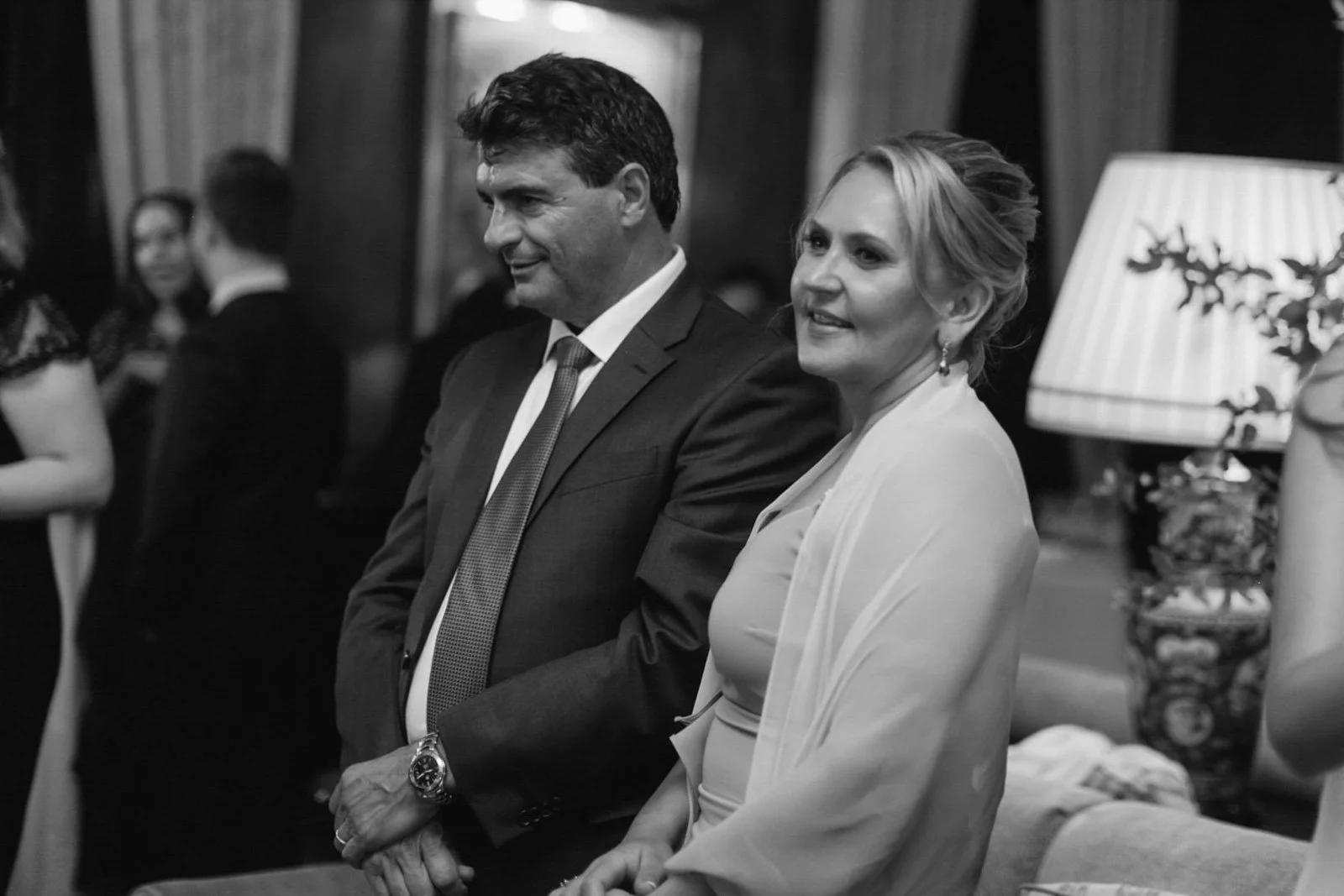 A black-and-white photo of a man and woman standing indoors, dressed formally, possibly at a social event or gathering.