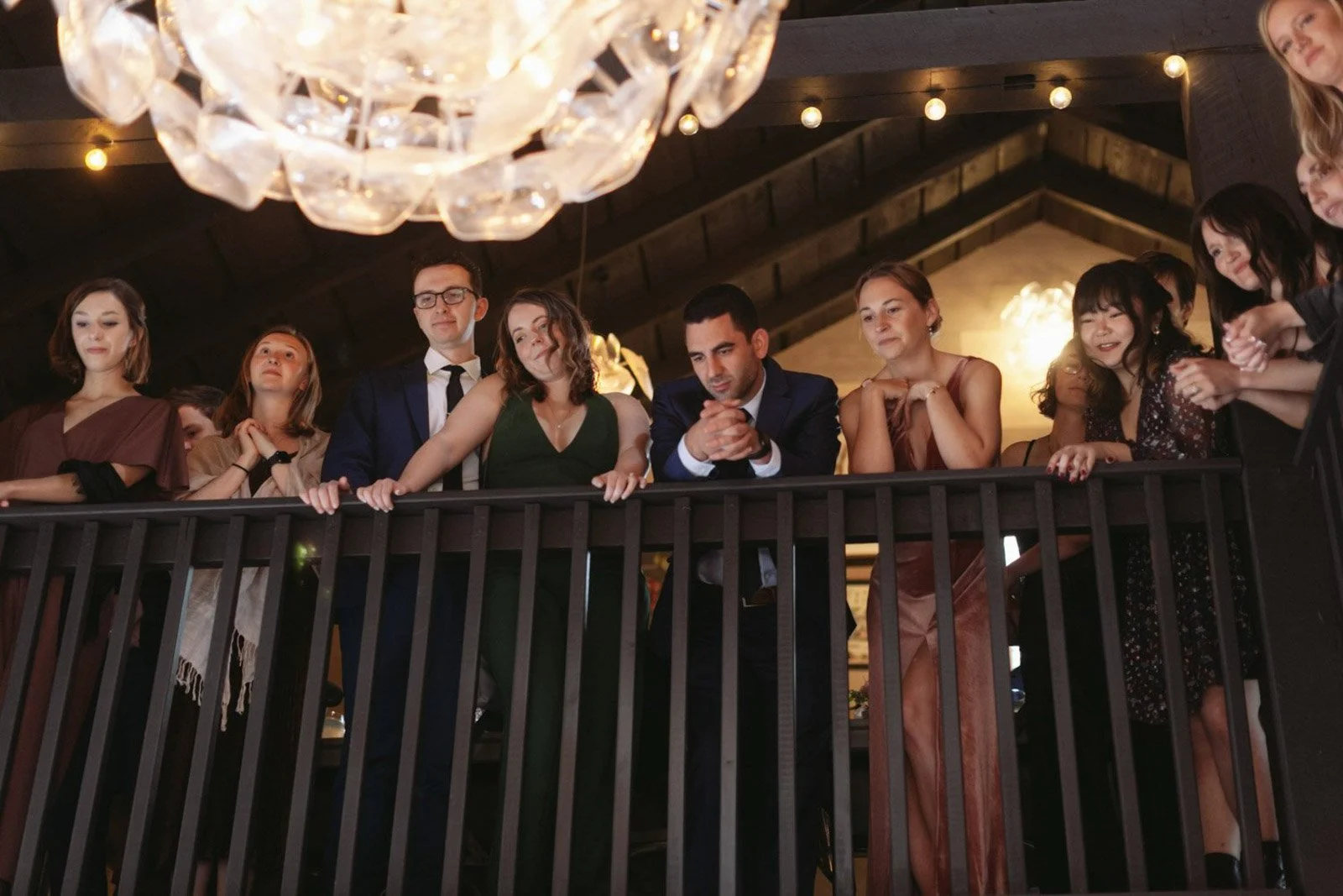 Group of people, mostly women, looking over a balcony at an indoor event, with a chandelier above.