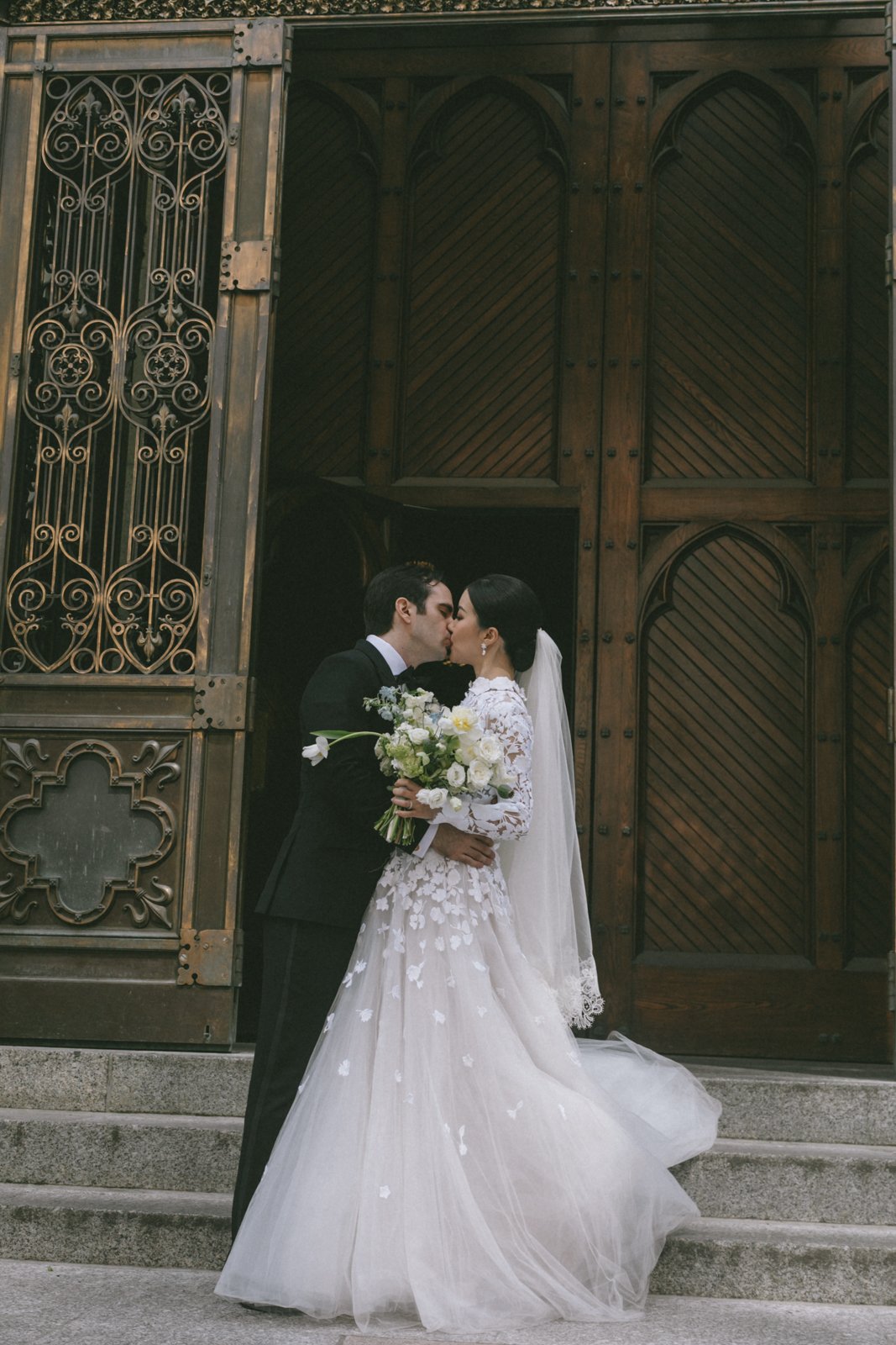 A bride and groom share a kiss on the steps outside a building with ornate wood and metal doors, the bride in a white wedding dress holding a bouquet, and the groom in a black tuxedo.