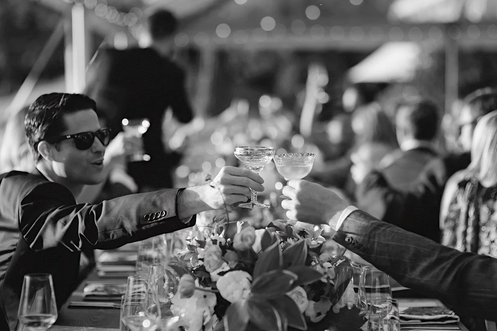 Two men in formal attire and sunglasses toast with cocktails at a dinner table decorated with flowers, during a celebration or event.
