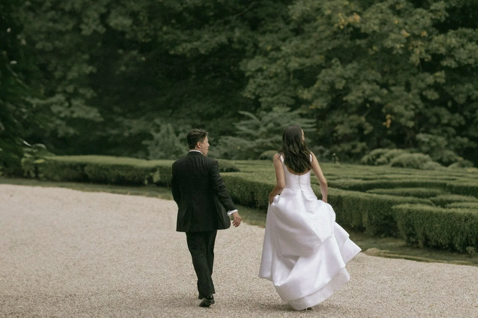 A man in a tuxedo and a woman in a white wedding dress walking together in a garden with neatly trimmed hedges and trees.
