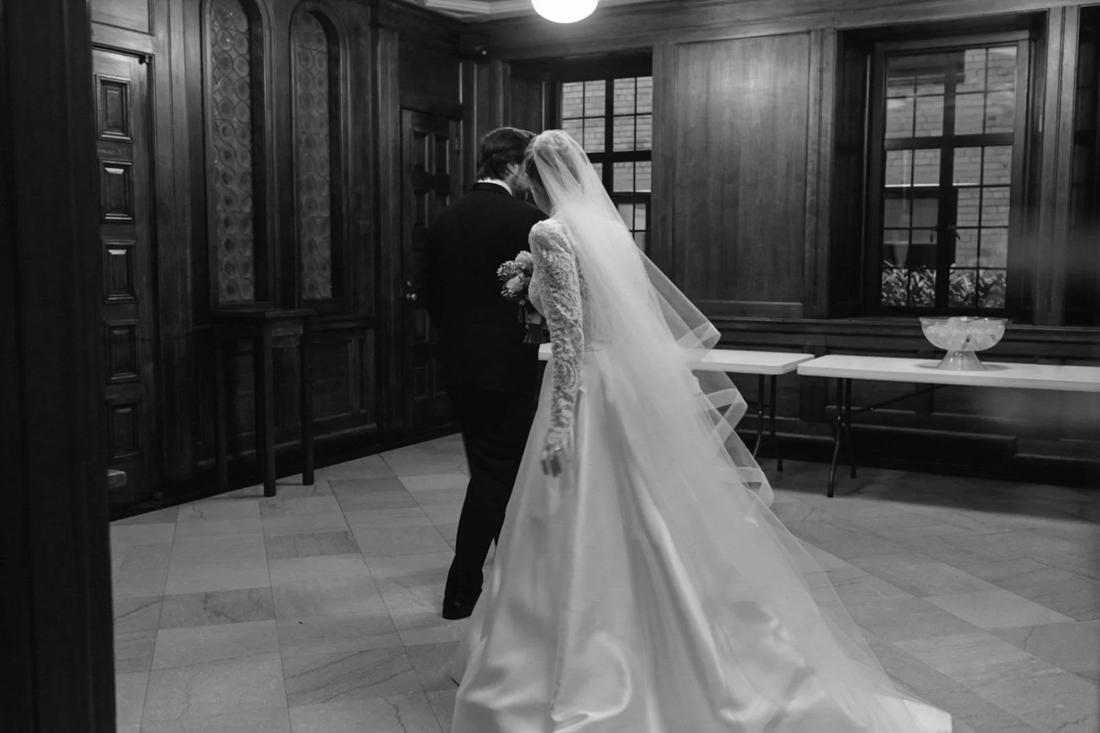 A bride and groom sharing a moment inside a wood-paneled room with large windows, the bride wearing a long wedding gown and veil, holding a bouquet, and the groom dressed in a suit.