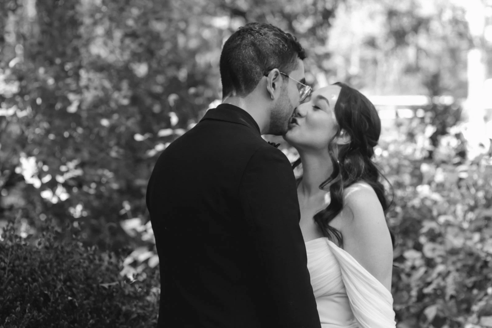 A black and white photo of a couple kissing outdoors, with trees and foliage in the background.