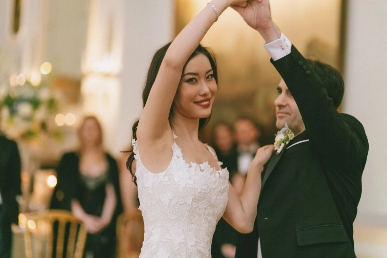 A bride and groom dance at their wedding reception, surrounded by blurred guests and elegant decorations.