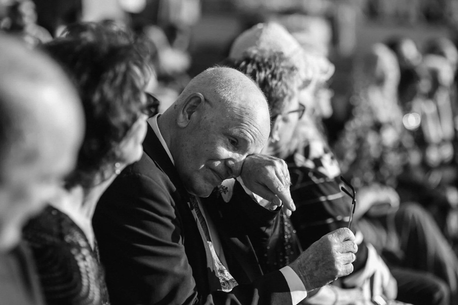 Black and white photo of elderly man with bald head sitting among a group of older people, looking down at a piece of paper or photograph, with others around him engaged in the same activity.