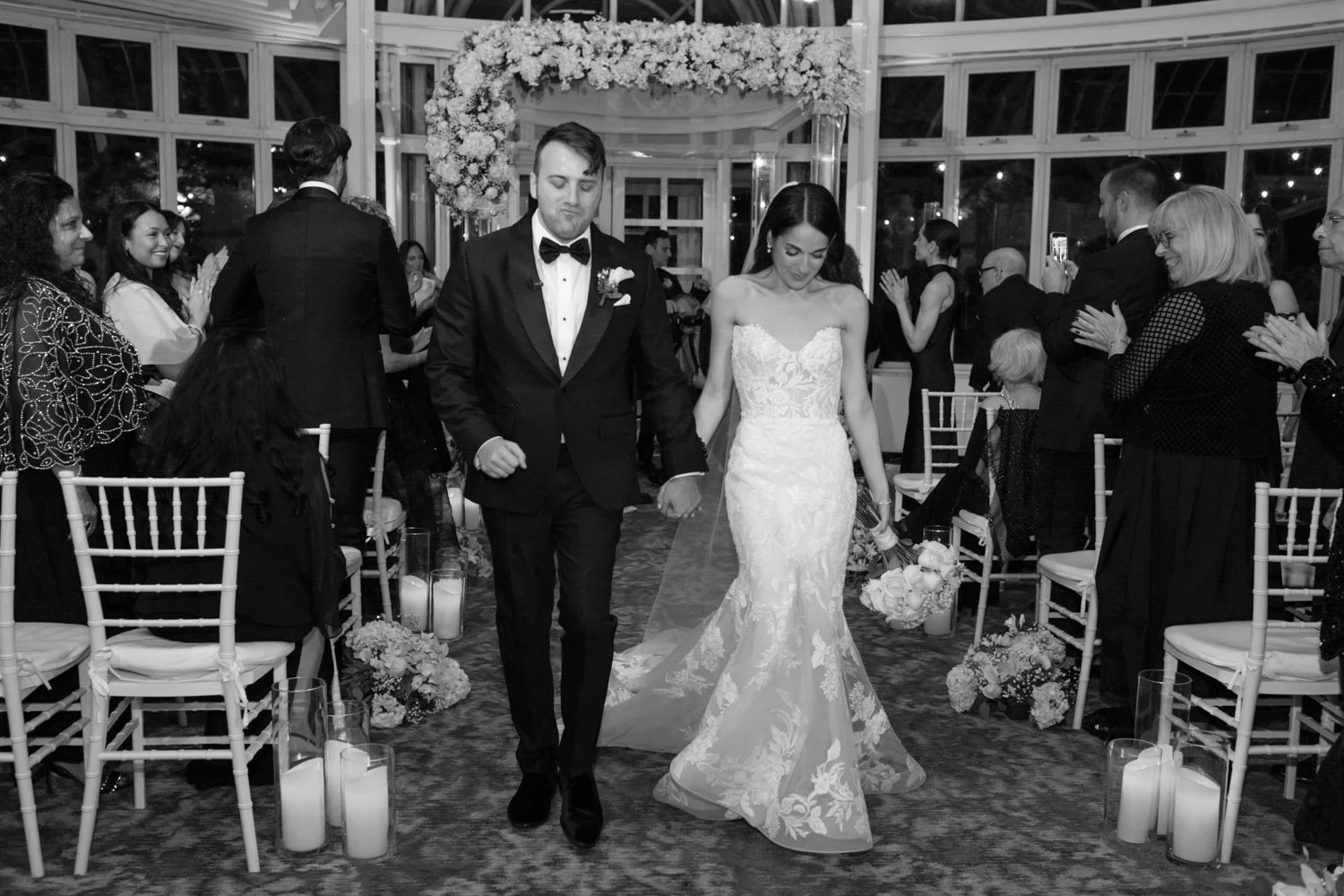 Black and white photo of a wedding ceremony showing a bride and groom holding hands and walking down the aisle, surrounded by seated guests clapping and smiling.