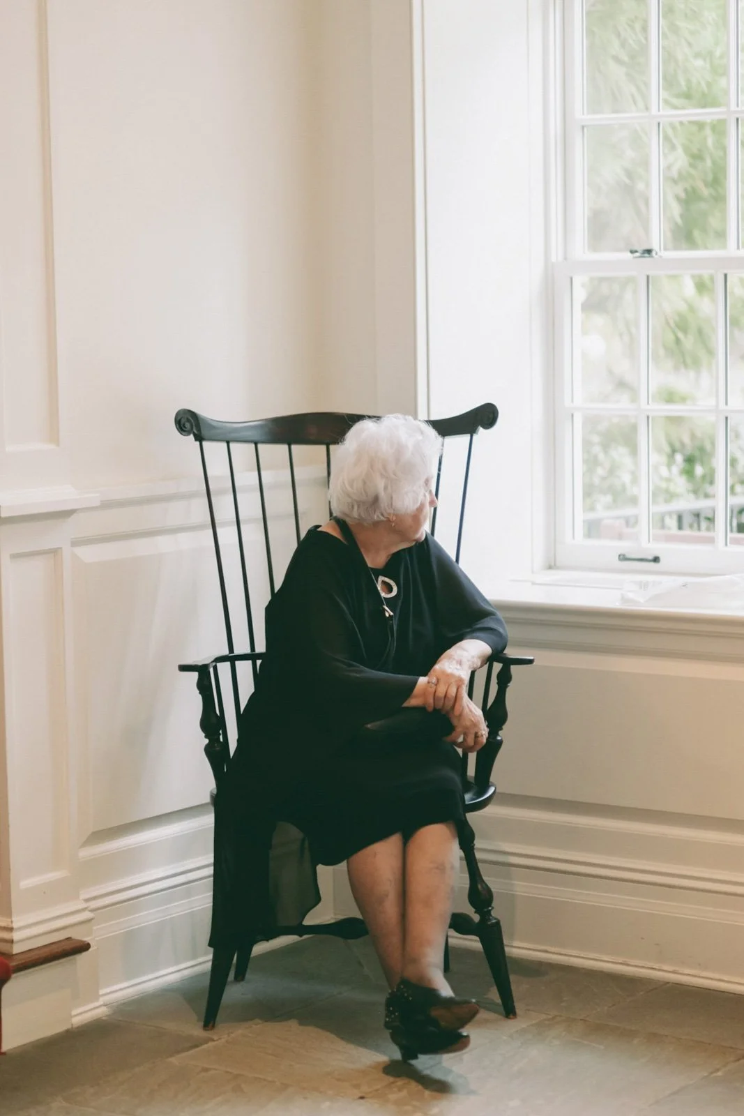 An elderly woman with white hair sitting in a black wooden chair near a window with a view of greenery outside. She is dressed in black and wearing jewelry, looking out the window.