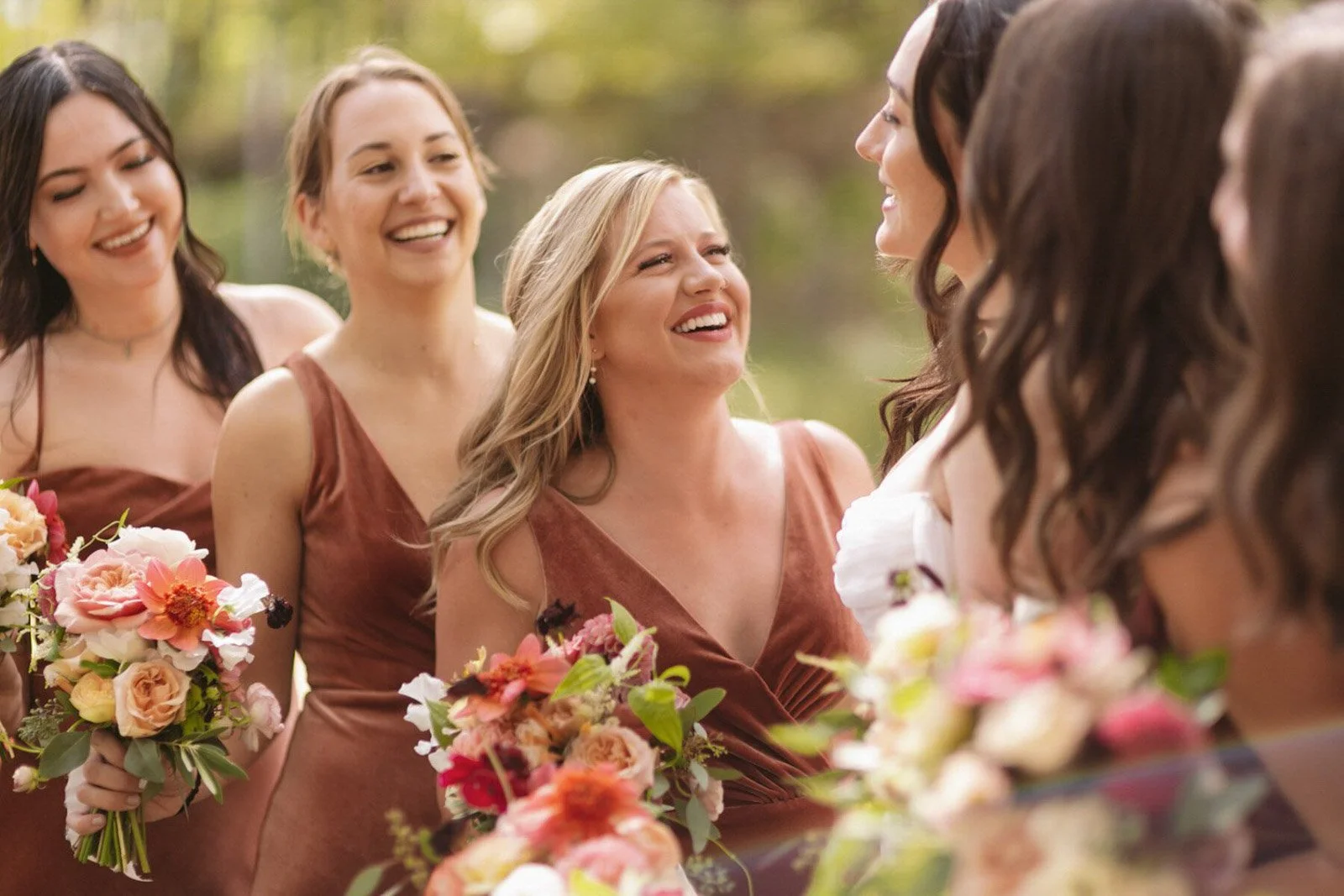 A group of women dressed in brown and white dresses holding bouquets of flowers, smiling and talking to each other outdoors during daytime.