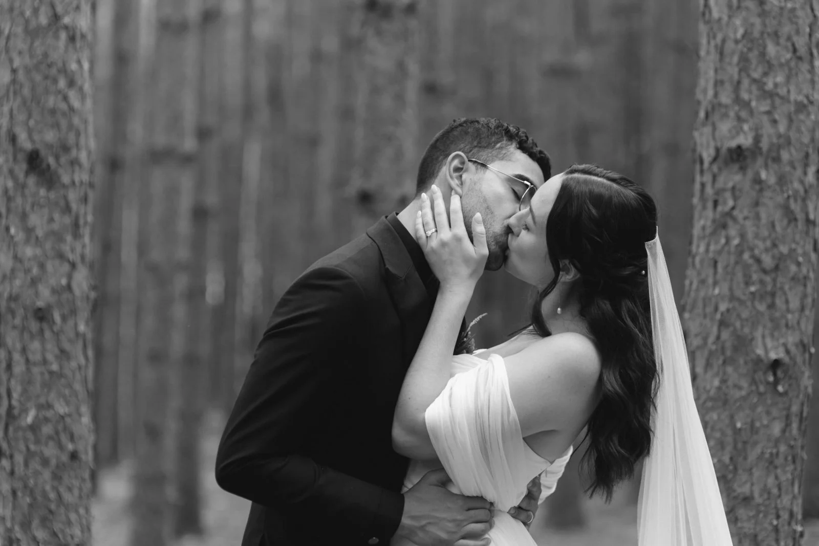Black and white photo of a bride and groom sharing a kiss outdoors, surrounded by trees.
