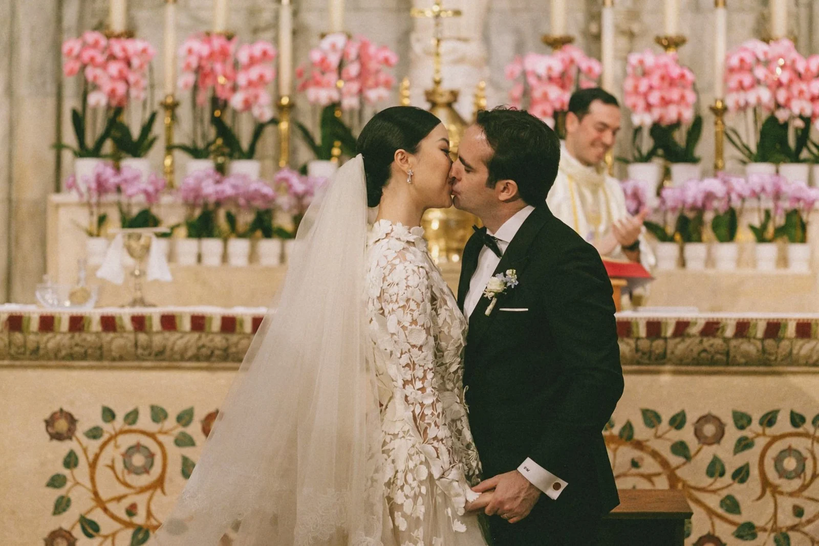 A bride and groom kissing during their wedding ceremony in a church, with a priest in the background and pink flowers decorating the altar.