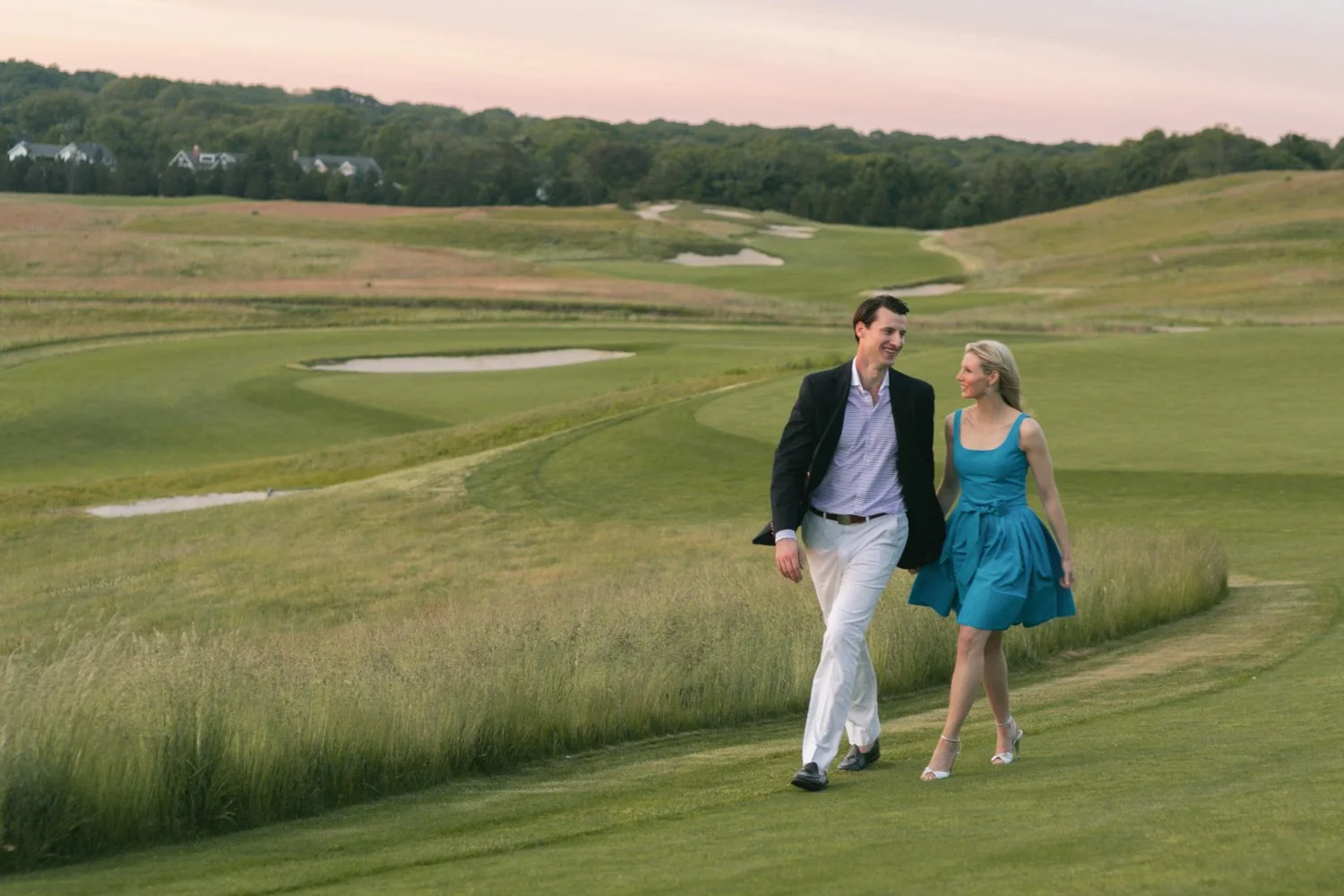 A couple walking and talking on a golf course, with lush green grass, sand traps, and distant trees in the background during sunset.