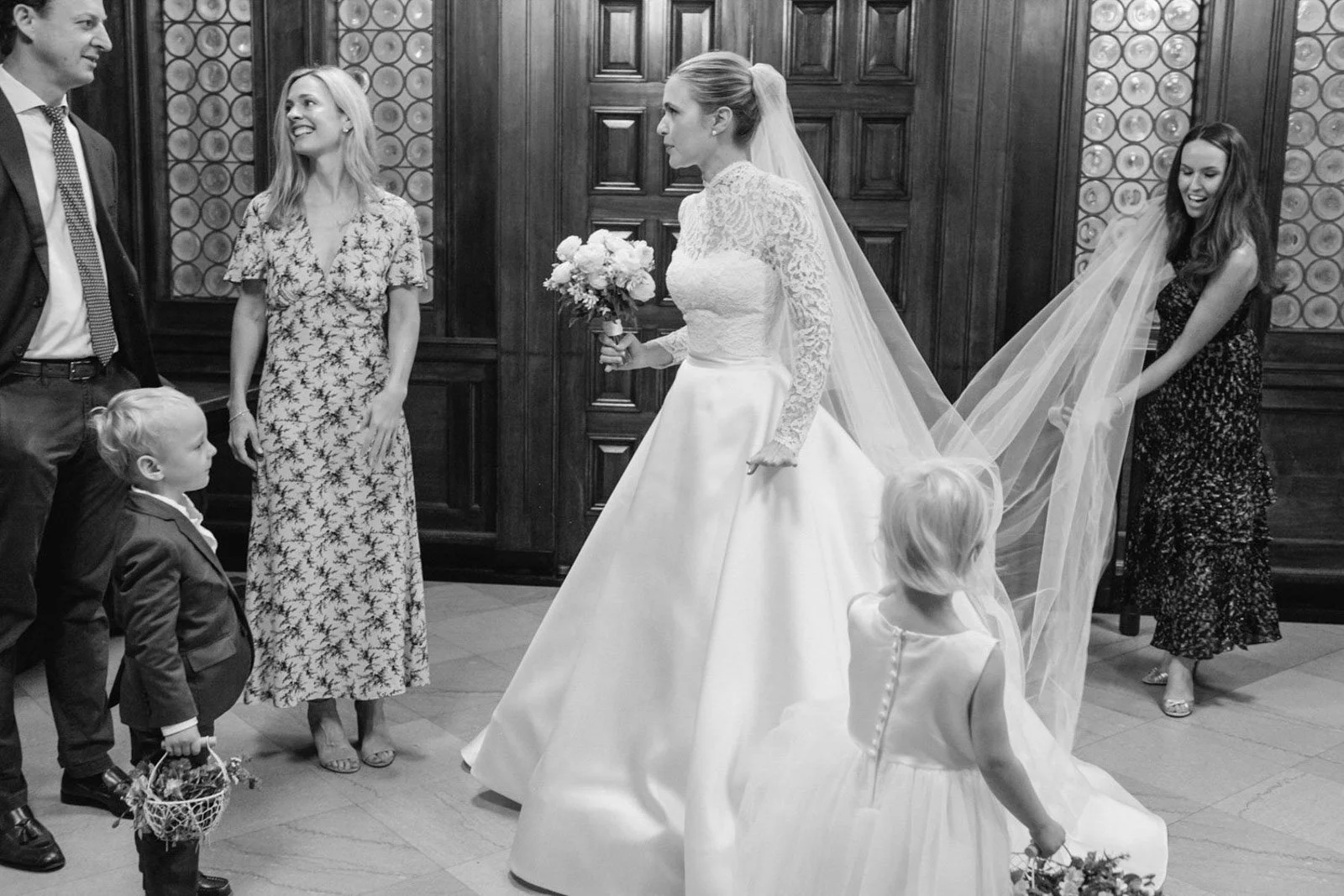 A bride in a lace and satin wedding gown holding a bouquet in a room with wood-paneled walls, surrounded by children and women in formal dresses, with one woman holding her wedding veil.