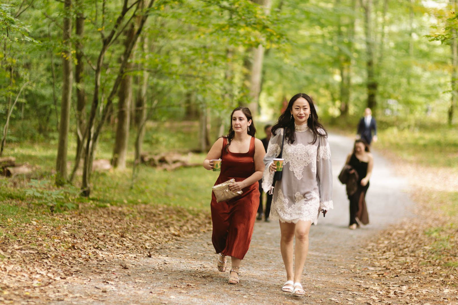 Four women walking on a forest trail, holding cups and wearing elegant dresses.