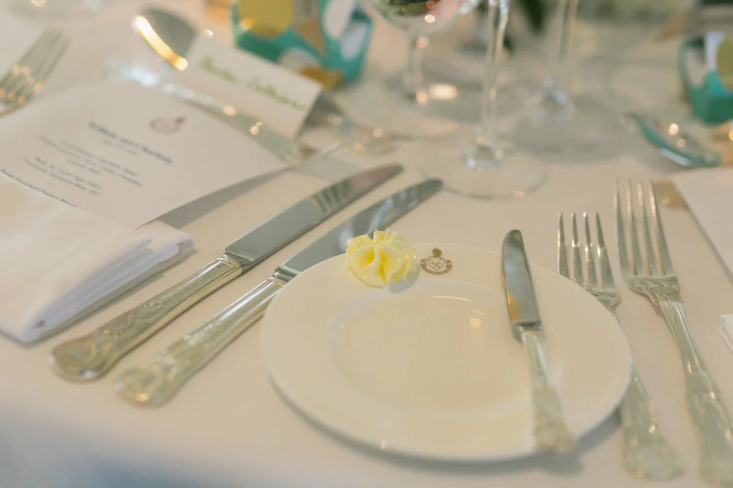 A formal dining table setup with silverware, a white plate with a small dollop of butter, a printed menu, and glasses, suggesting a banquet or wedding reception setting.