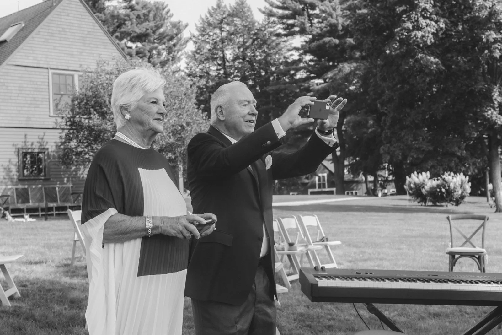 Two elderly people, a woman and a man, standing outdoors in a garden or park. The man is taking a selfie or recording a video with his phone, while the woman watches and smiles. There is a keyboard instrument in front of them and several chairs in th