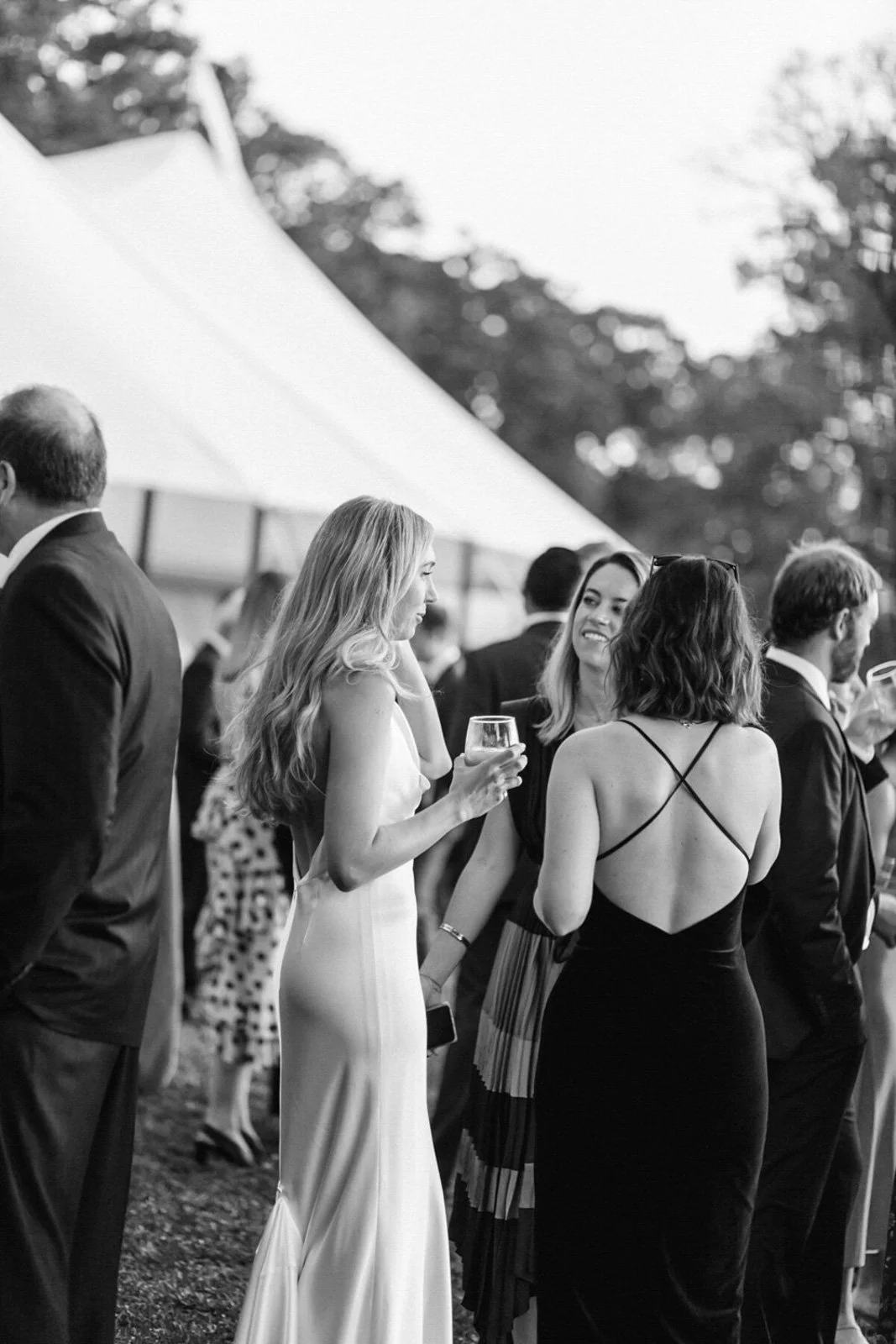 People in formal attire at an outdoor event, socializing and holding drinks, under a large tent.