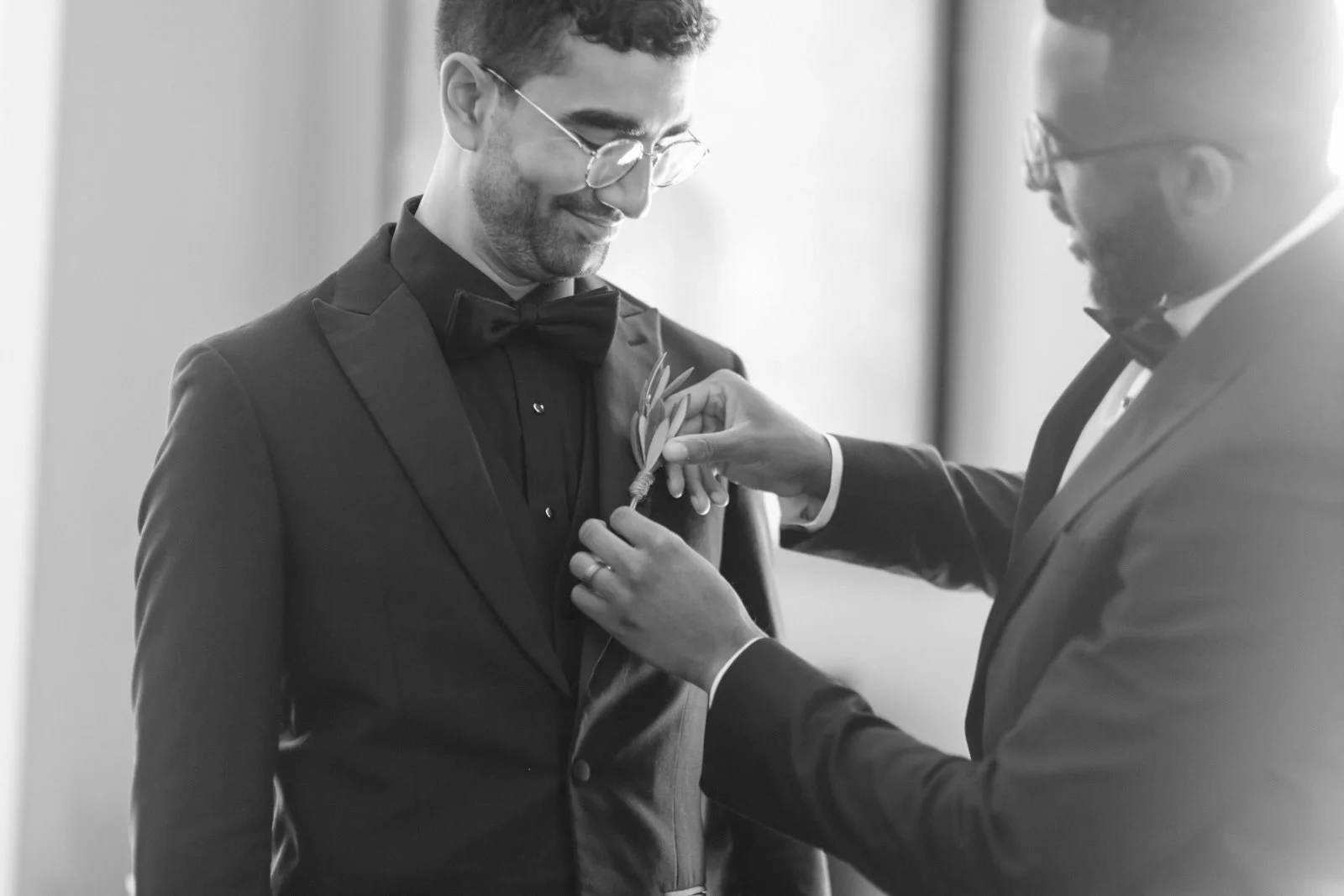 Two men in tuxedos, one pinning a boutonniere on the other's lapel during a wedding or formal event.