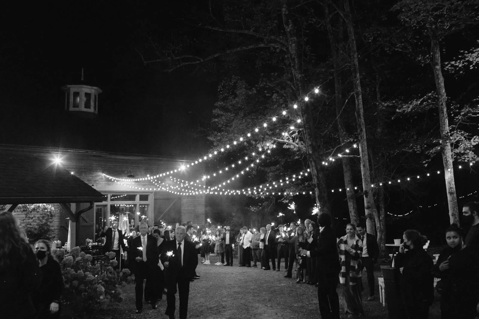 People dressed in formal attire attending nighttime outdoor event with string lights hanging overhead in a wooded area.
