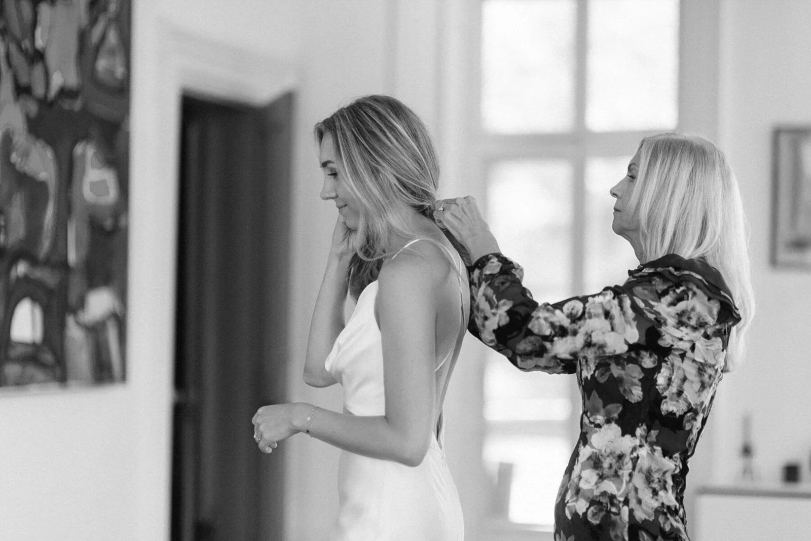 An older woman with long, light-colored hair helping a young woman with long hair, dressed in a white dress, with her hair in a ponytail, on a bridal getting-ready process in a bright room.