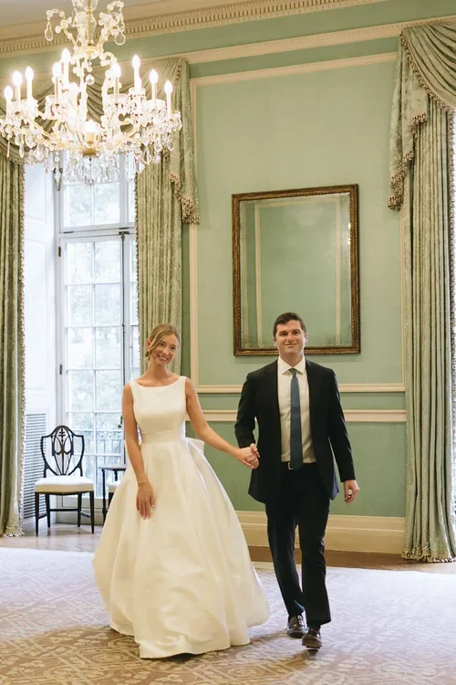 A bride in a white wedding dress and a groom in a suit walking hand in hand in an elegantly decorated room with a chandelier, framed mirror, and large windows.