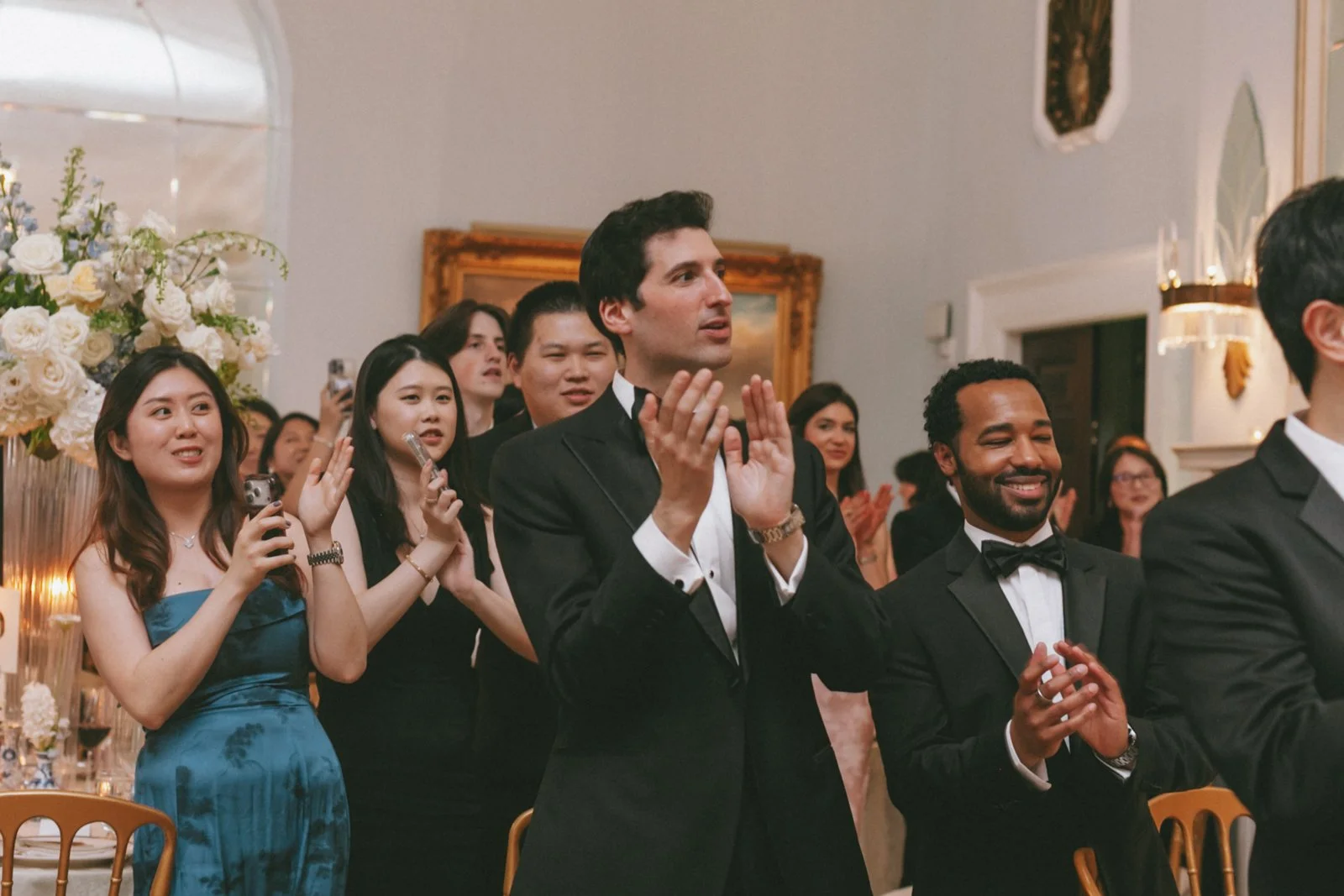 People clapping and smiling at a formal event, with a focus on a man in a tuxedo clapping and a woman in a blue dress holding a phone.
