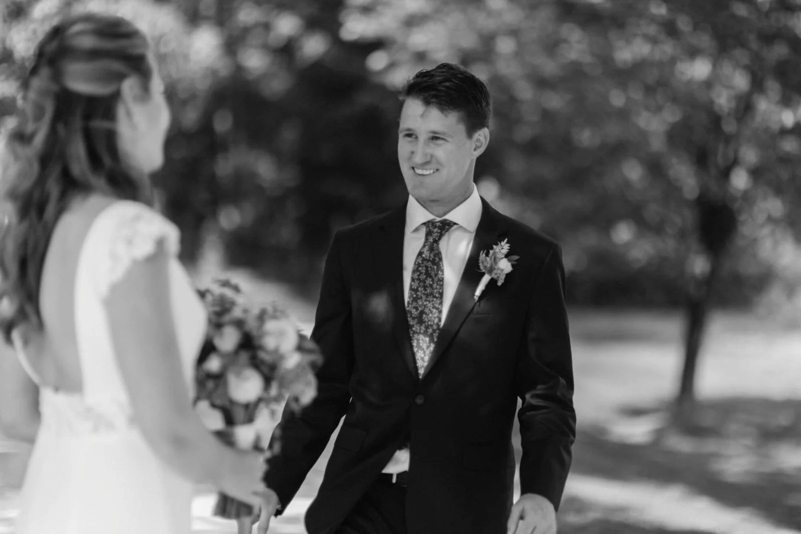 A black-and-white photograph of a groom smiling as he holds the hand of a bride, who is holding a bouquet, during a wedding ceremony outdoors.