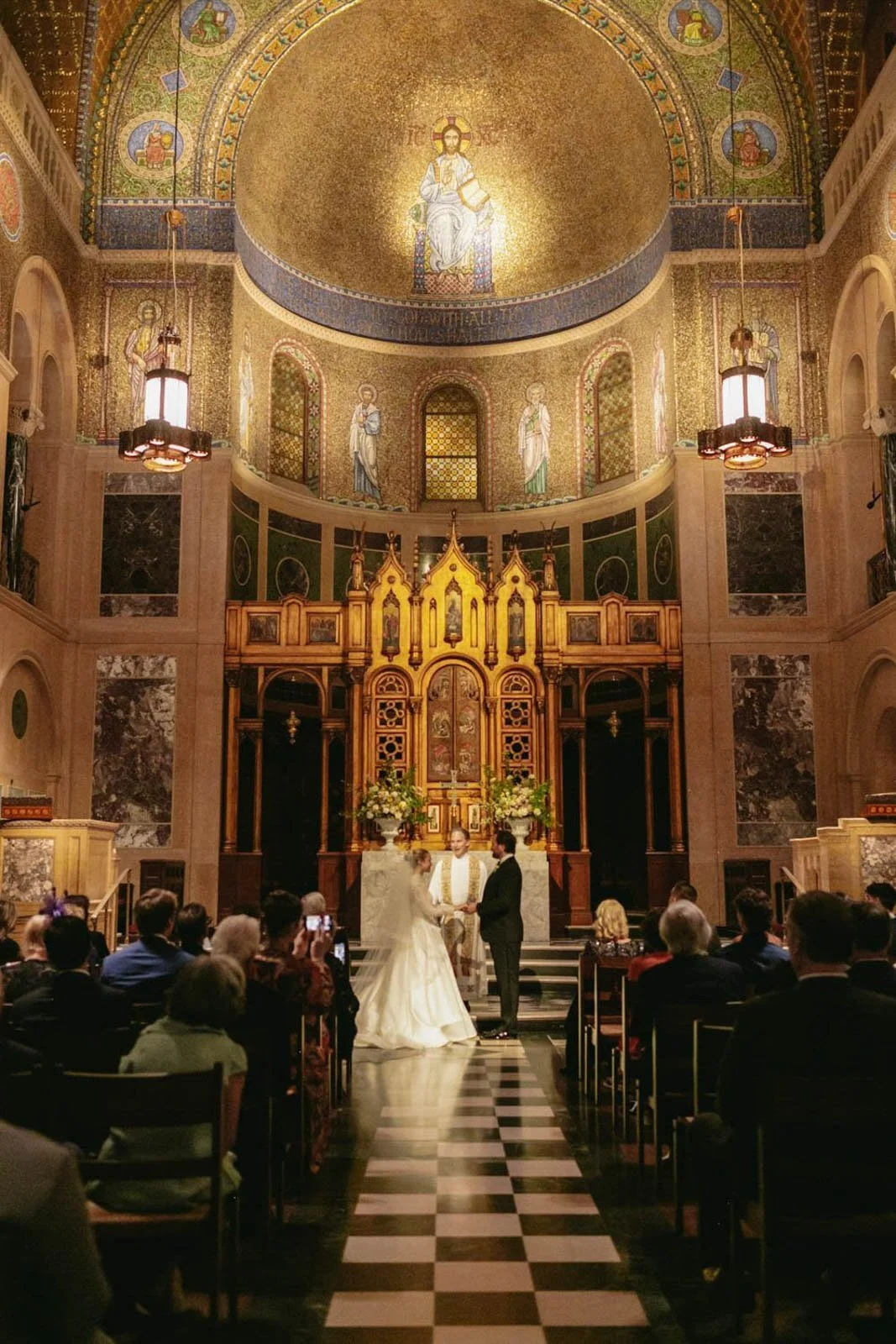 A wedding ceremony taking place inside a church with an ornate altar and religious artwork decorated with gold mosaics. Bride and groom are holding hands in front of a priest, with guests seated on either side of the aisle.