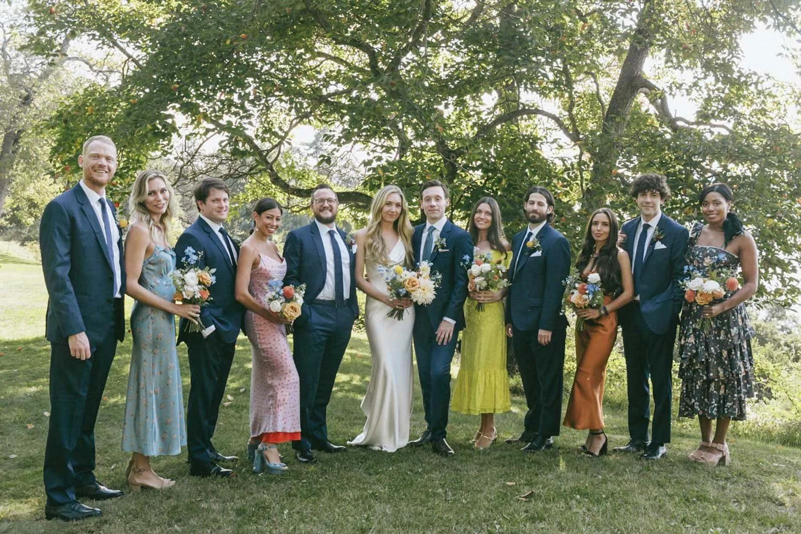 Group of people dressed in formal attire, holding bouquets, standing outdoors in front of a large tree during daytime.