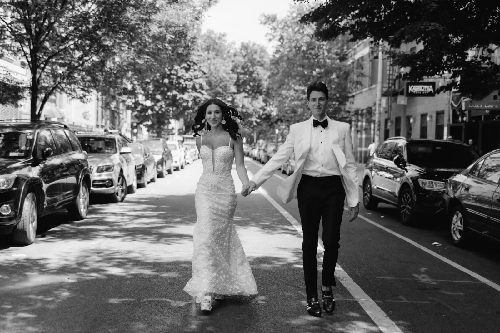 A couple in wedding attire walking hand in hand on a city street, with parked cars and trees lining the streets in the background.