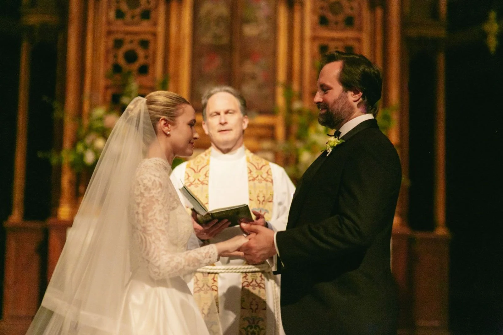 A bride and groom holding hands during their wedding ceremony, standing before a priest in a church with ornate wooden decor.
