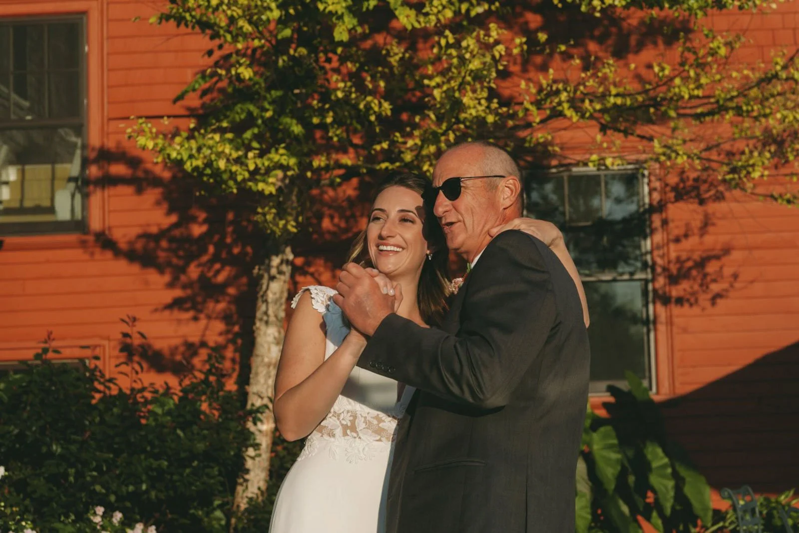 A bride dancing with an older man outdoors during sunset, smiling, with a red wooden building and green foliage in the background.