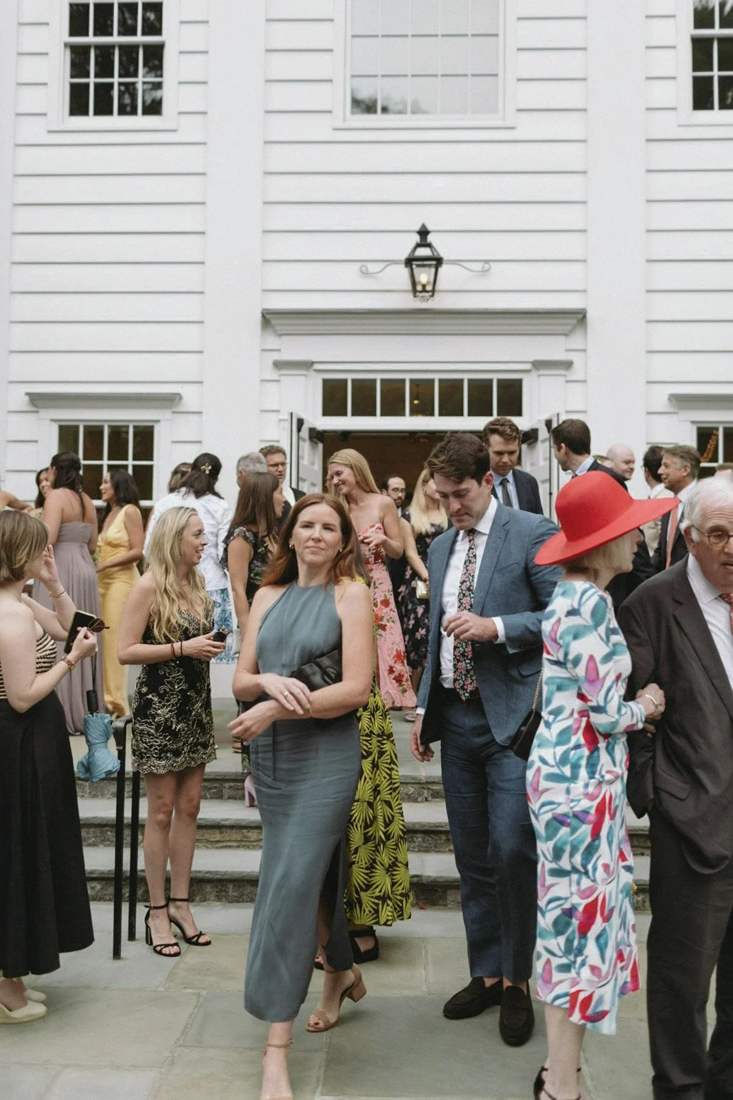 Group of people dressed in formal and semi-formal attire socializing outside Stanwich Church in Greenwich, CT, with steps, large windows, and a lantern above the entrance.