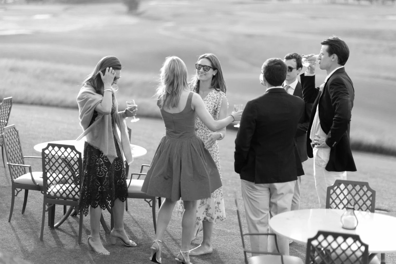 Group of six people at an outdoor gathering, socializing and drinking wine, with tables and chairs around them.