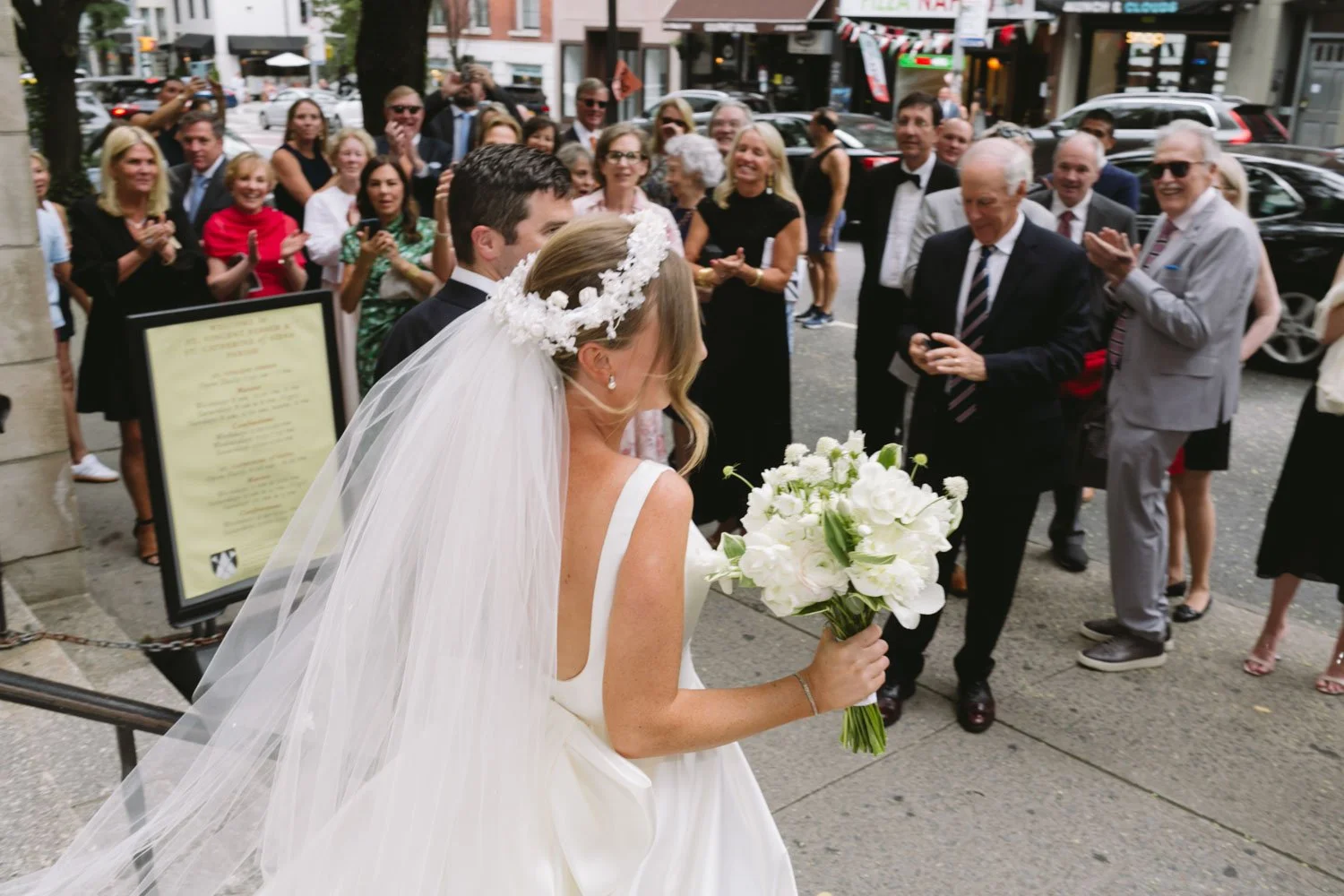 Bride in a white wedding dress with a floral headpiece and veil holding a bouquet of white flowers, walking on a city sidewalk surrounded by celebrating guests.